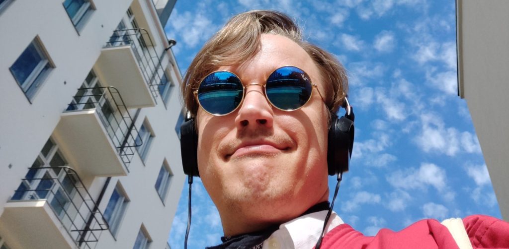 Punatakkinen, aurinkolasipäinen mies katsoo kameraan kohti alaviistoa.